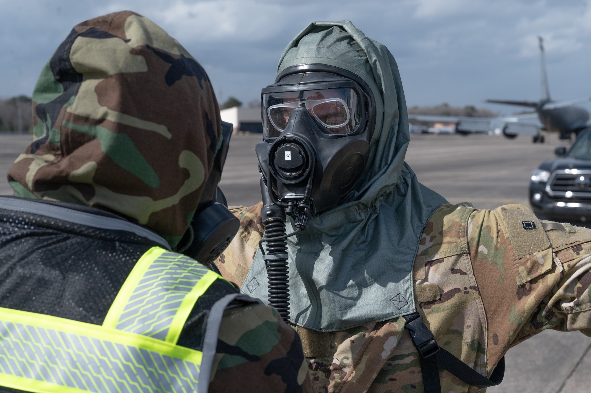 An aircrew member wearing a chemical protection suit prepares to have his hood removed in a contamination control area.