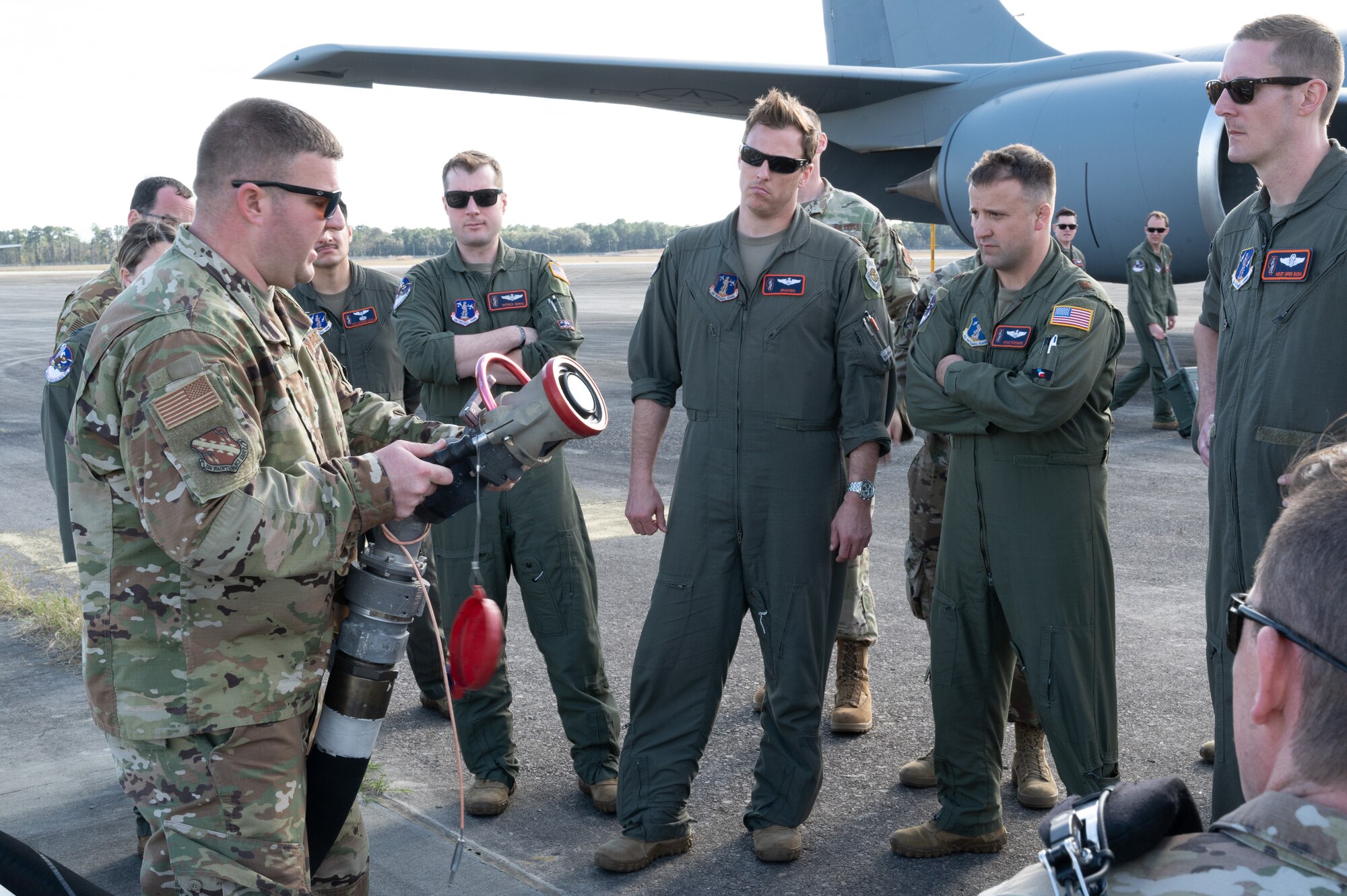 A group of Air Force pilots stand watching as a maintenance personnel holds up a aircraft fuel hose and explains how to use it.