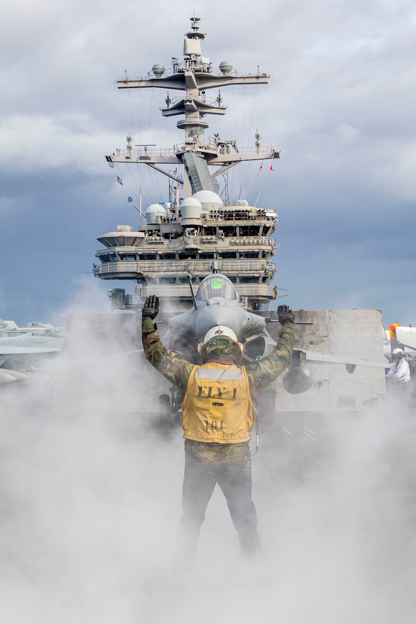 (Nov. 23, 2022) Aviation Boatswain’s Mate (Handling) 3rd Class Roberto Cerdas assigned to the Nimitz-class aircraft carrier USS George H.W. Bush (CVN 77), directs a French Rafale fighter jet onto the catapult during multi-carrier operations between the George H.W. Bush Carrier Strike Group (CSG), Charles de Gaulle CSG, and the Italian Cavour CSG Nov. 23, 2022. The George H.W. Bush CSG is on a scheduled deployment in the U.S. Naval Forces Europe area of operations, employed by U.S. Sixth Fleet to defend U.S., allied, and partner interests.
