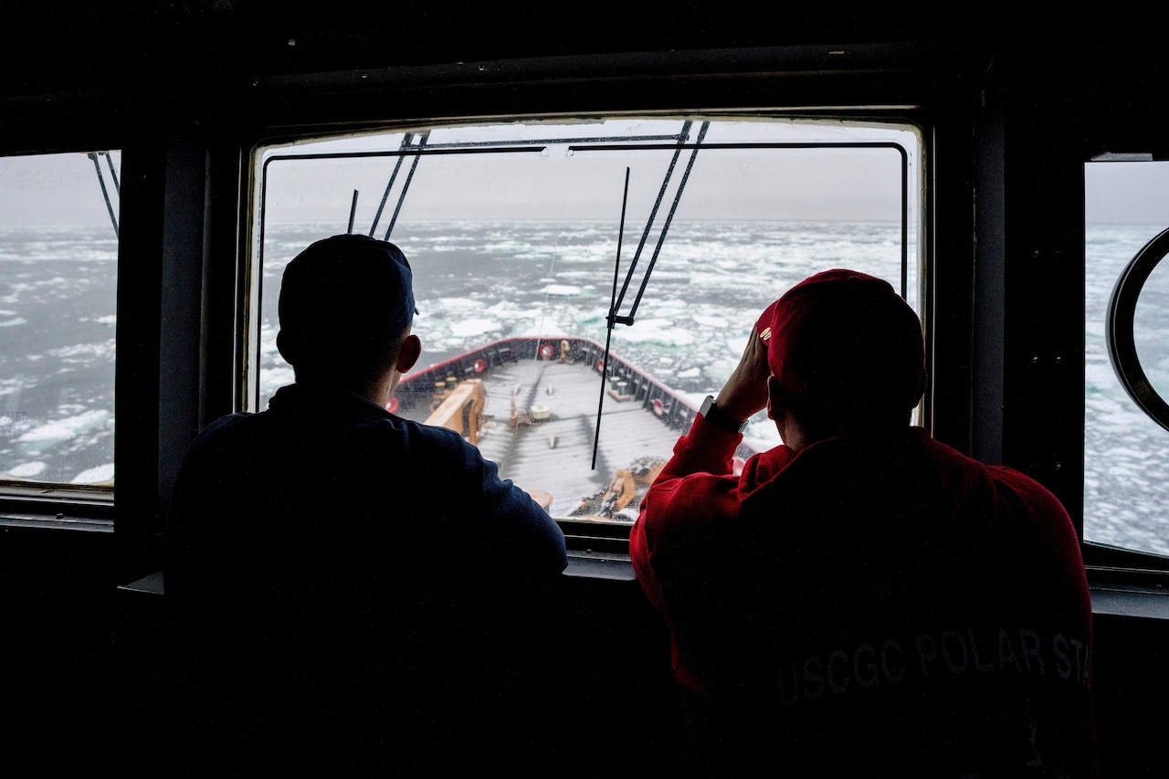 Two Coast Guardsmen look out from a window at the bow of a cutter in icy waters.