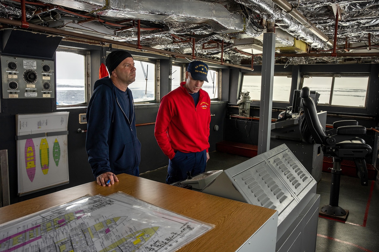 Two  Coast Guardsmen stand in an interior space on a cutter.
