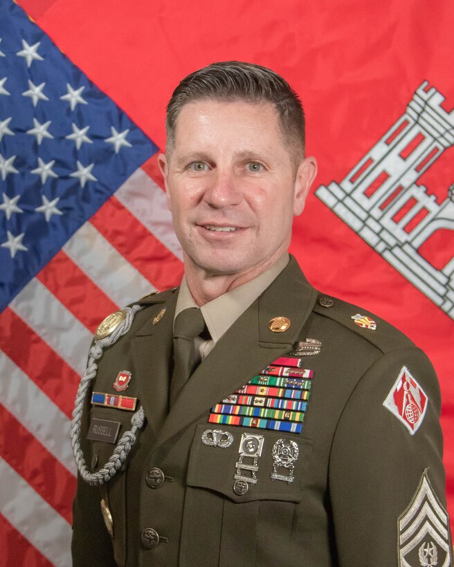 Male army soldier in dress uniform standing in front of US and USACE flags.