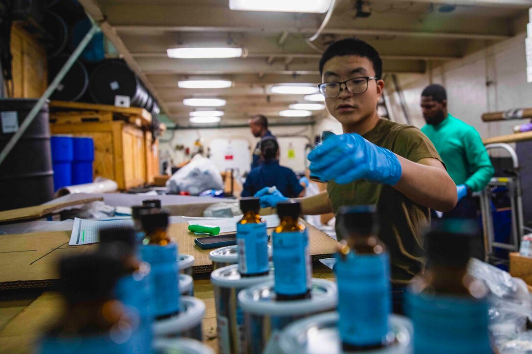 A sailor wearing gloves reaches for a small bottle on a table.