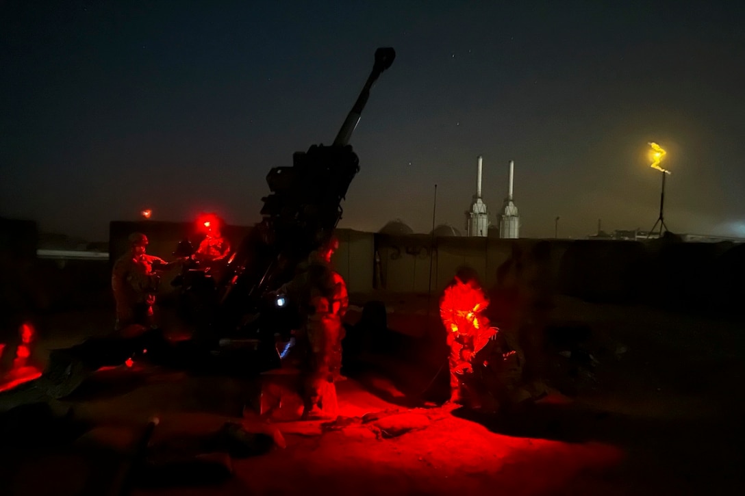 Soldiers stand next to a howitzer at night as red lights shine on them.