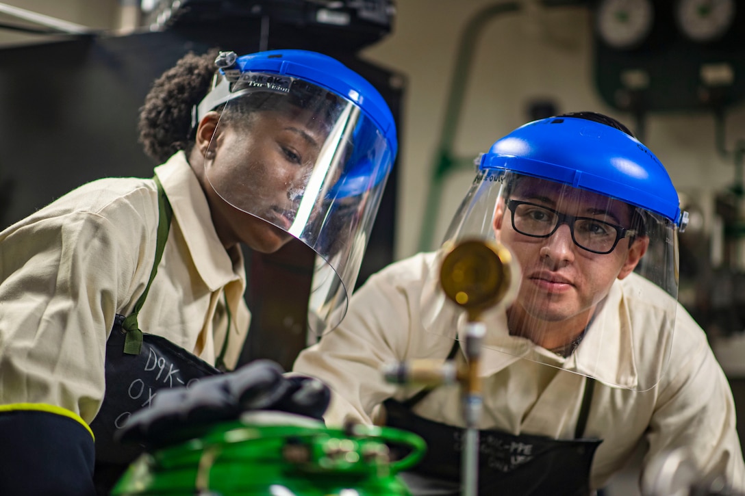 Two sailors wearing protective gear look at an object in a lab aboard a ship.