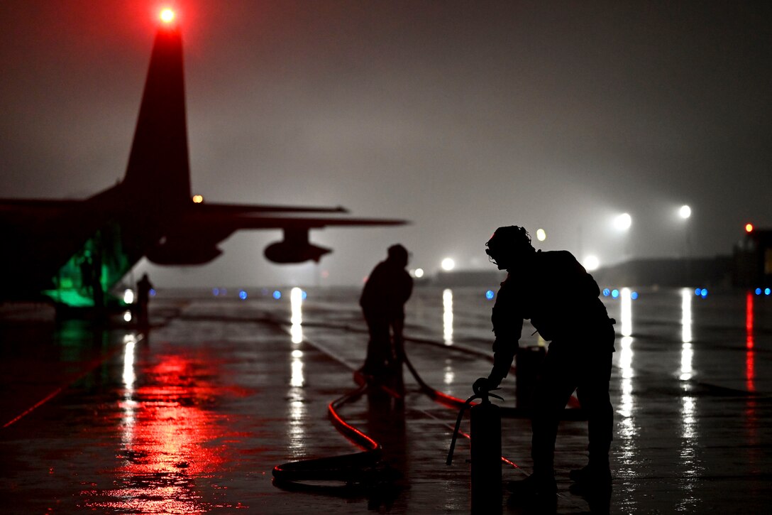 Two airmen hold fuel hoses near aircraft in the dark illuminated by red, blue, green and white lights.