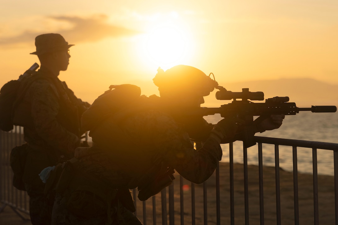 A Marine aims a weapon as a fellow Marine watches under a sunlit sky.