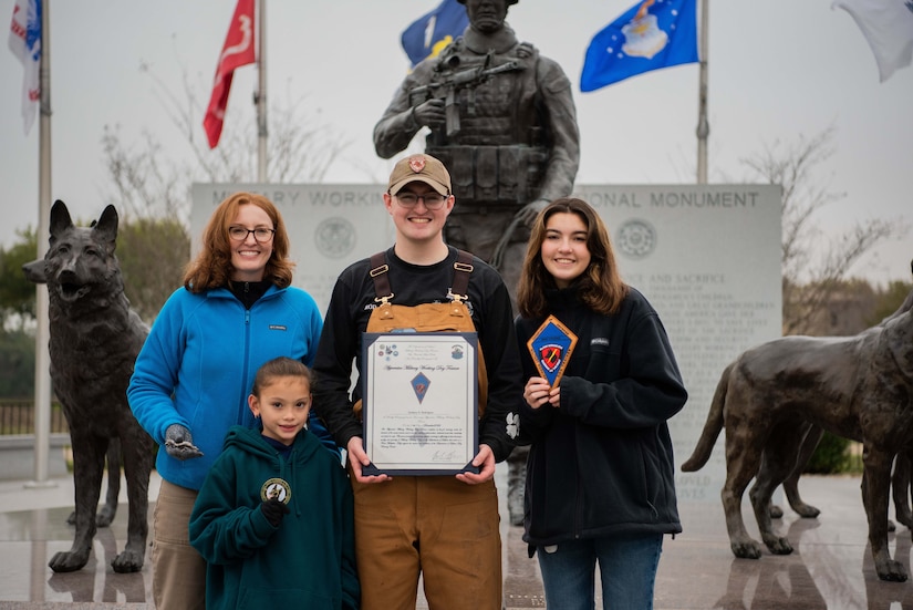 A young man holds a certificate against his chest and smiles for the camera. An older woman stands to his right, and a younger woman stands to his left. In front of the group stands a young girl.