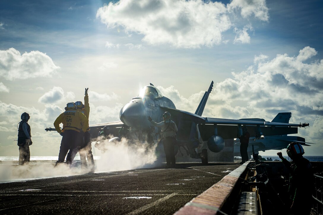 Several sailors stand amid smoke on a ship's flight deck and signal toward an aircraft with a cloudy and blue sky background.