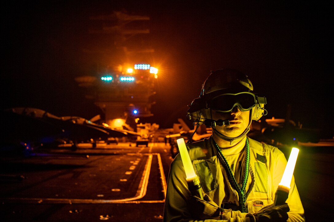 A sailor holds up two lit wands at night on the deck of a ship with lights on the tower in the background.