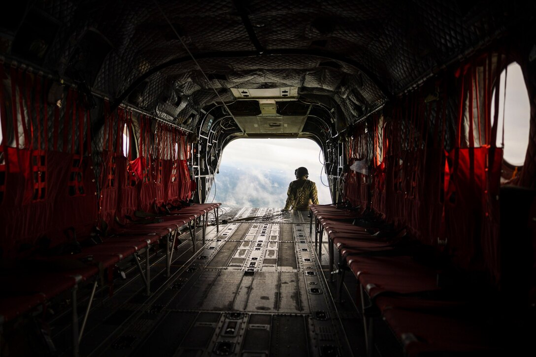 A soldier its at the back of an airborne aircraft looking out over land.