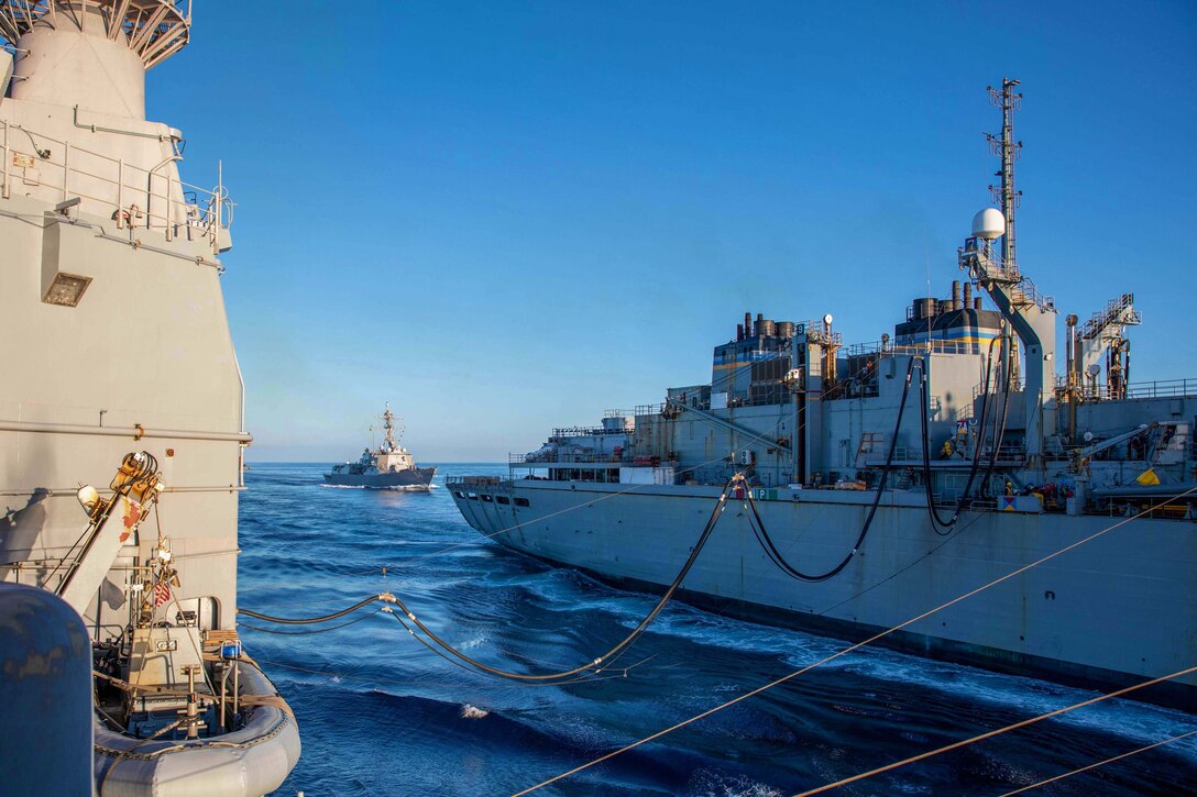 Two ships sail next to each other connected by a hose as another ship travels nearby.