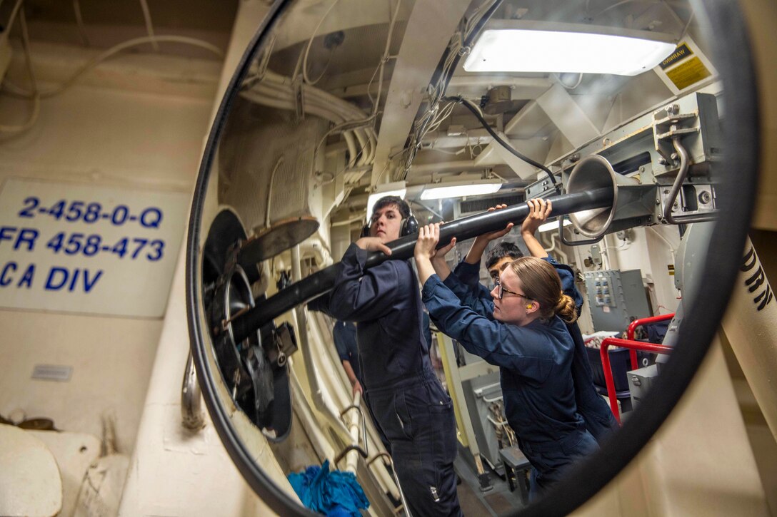 Sailors hold a cable in the air in a maintenance room as seen through the reflection of a mirror.