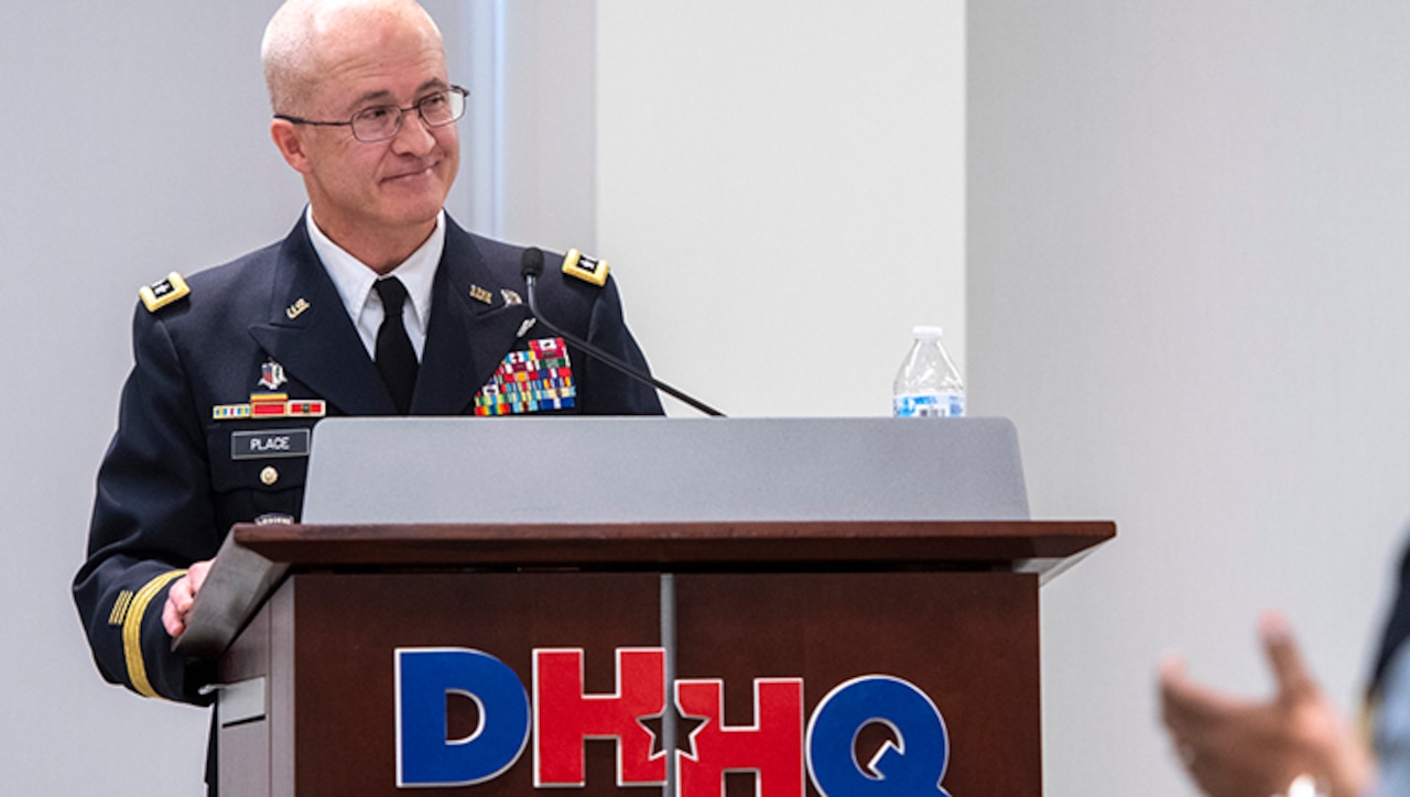 A military service member smiles while standing behind a lectern.