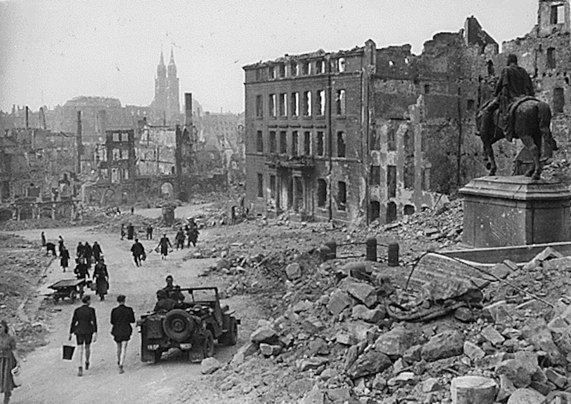 A vehicle travels and people walk through a city in ruins.