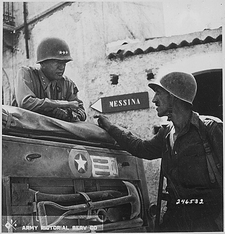 A man talks to another man leaning on a vehicle.