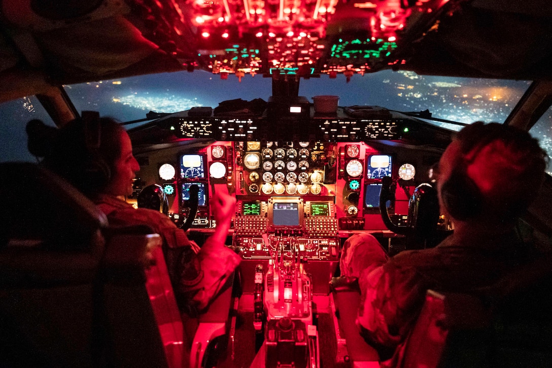 Two airmen talk while sitting in the cockpit of an airborne aircraft illuminated by colorful lights.