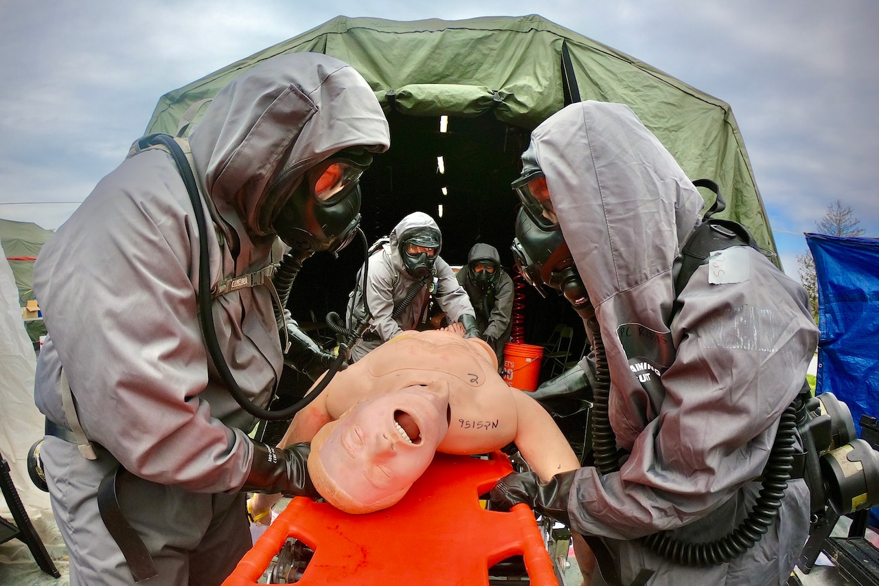 Four soldiers wearing hazmat suits work on a dummy that’s on a stretcher.