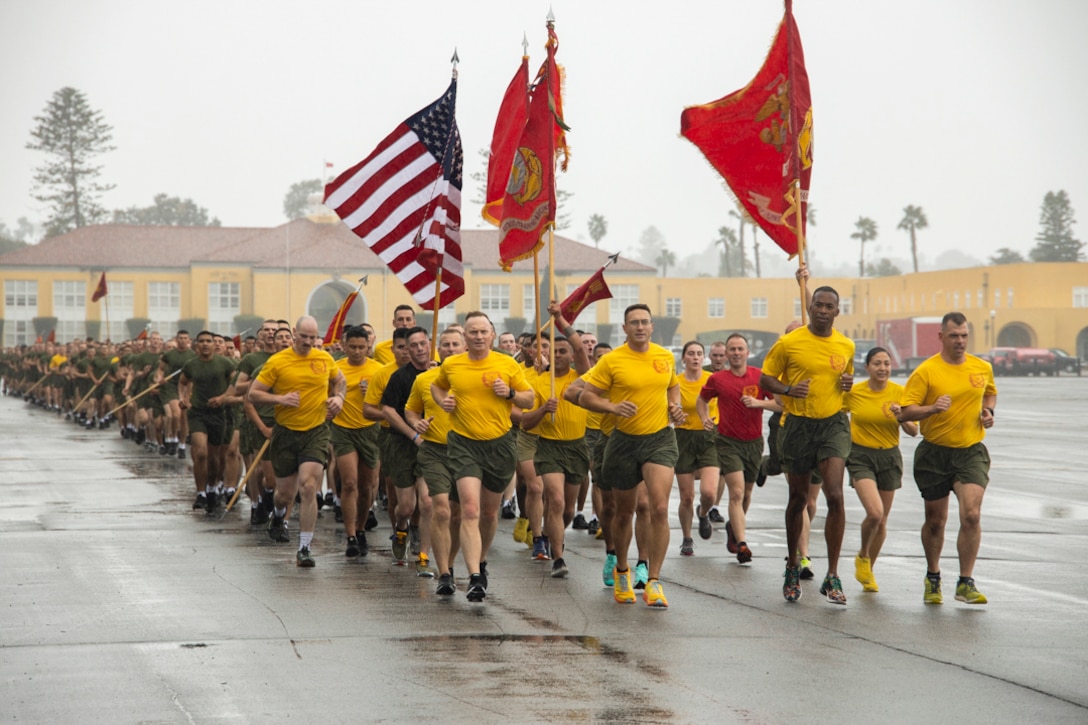 A large group of Marines run while holding flags on a rainy day.