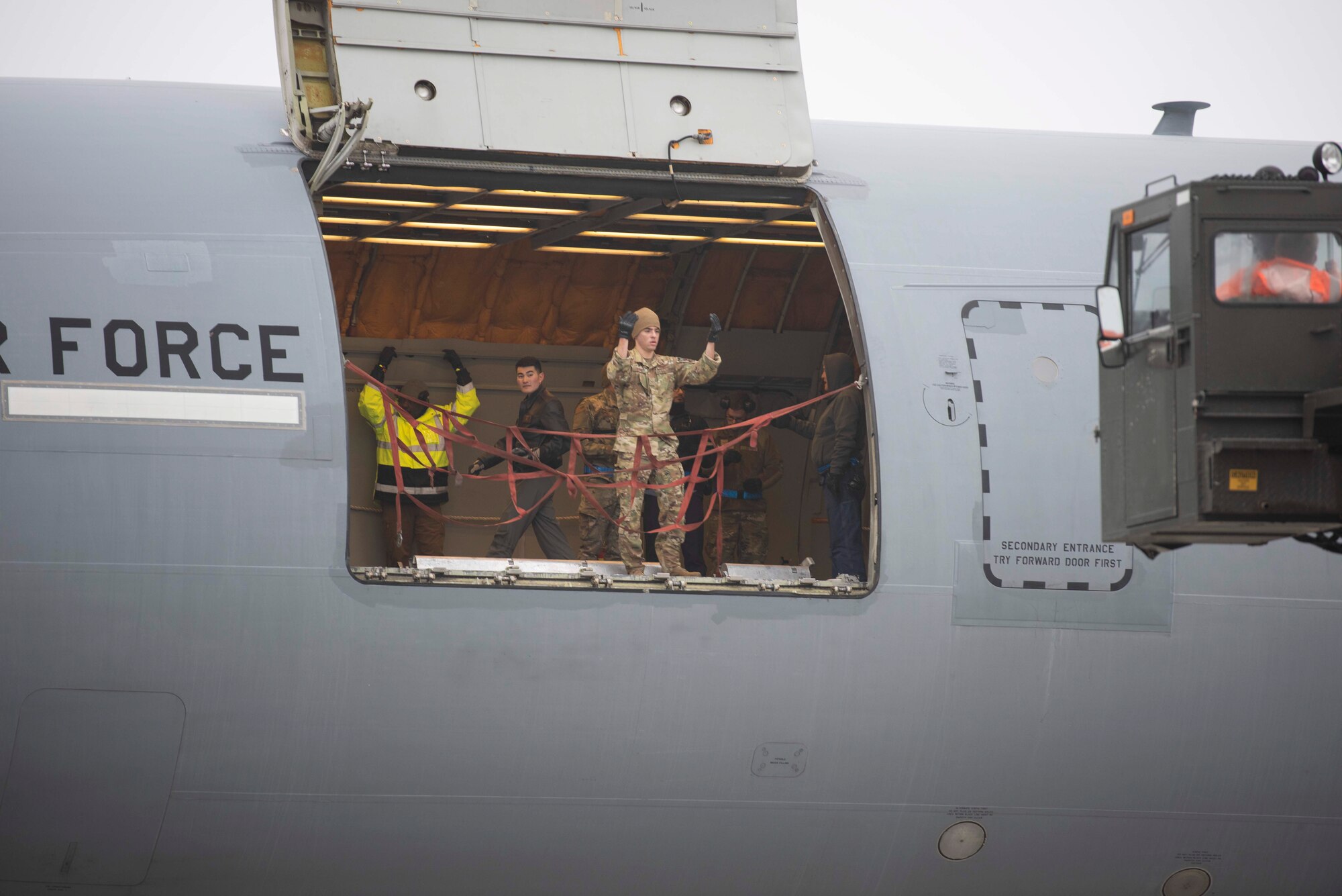 Military members stand near the cargo loading door of a large military aircraft giving signals to an approaching large military transport vehicle.