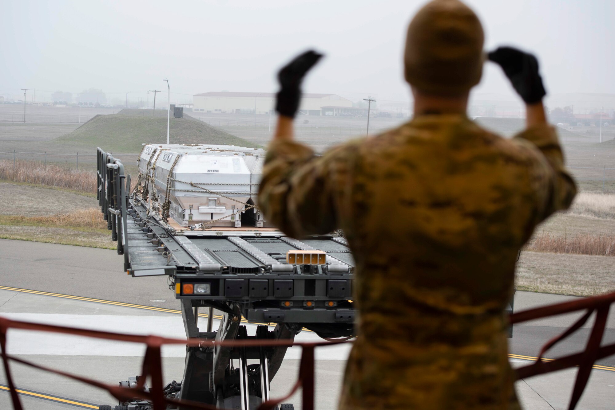A military member stands inside a large military aircraft while a large military transport vehicle is driven towards them to be loaded on to the aircraft.