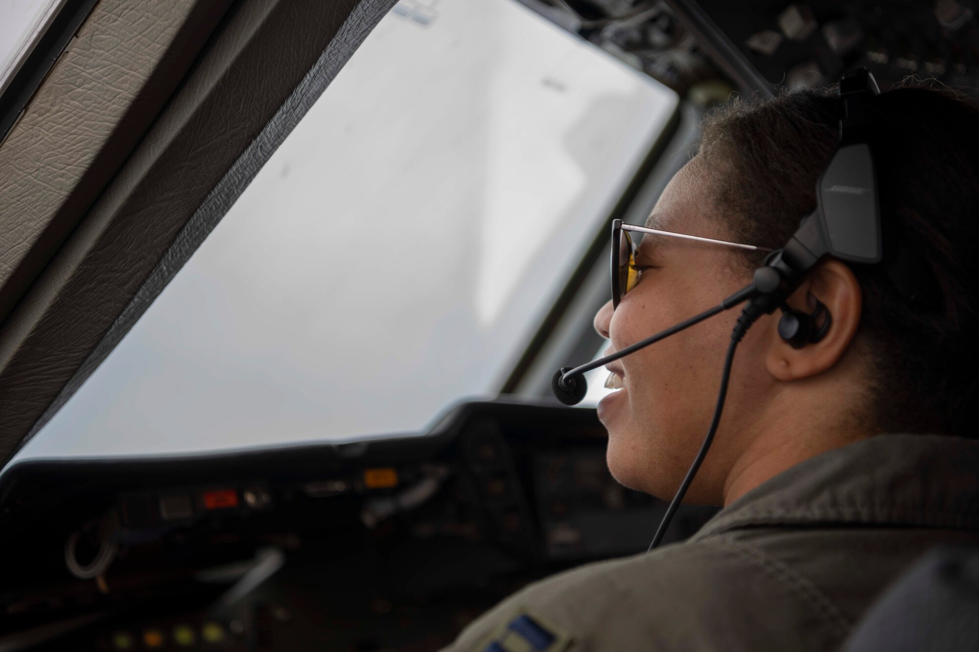 A military member wearing a headset device for communication smiles as they sit in a chair on the flight deck of a large military aircraft.