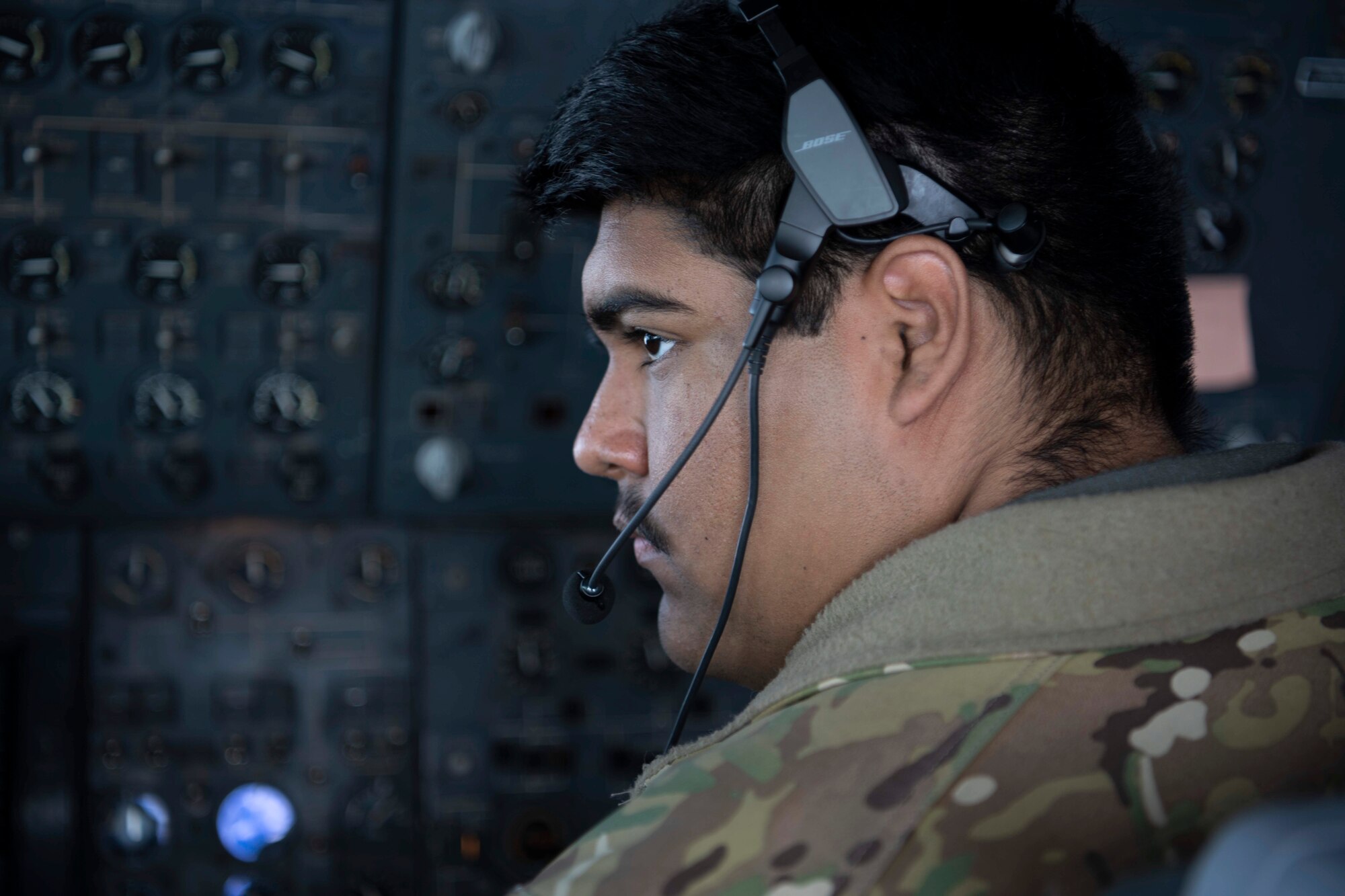 A military member wearing a headset for communications sits in a chair next to a large panel covered in buttons and switches for various operations on a large military aircraft.