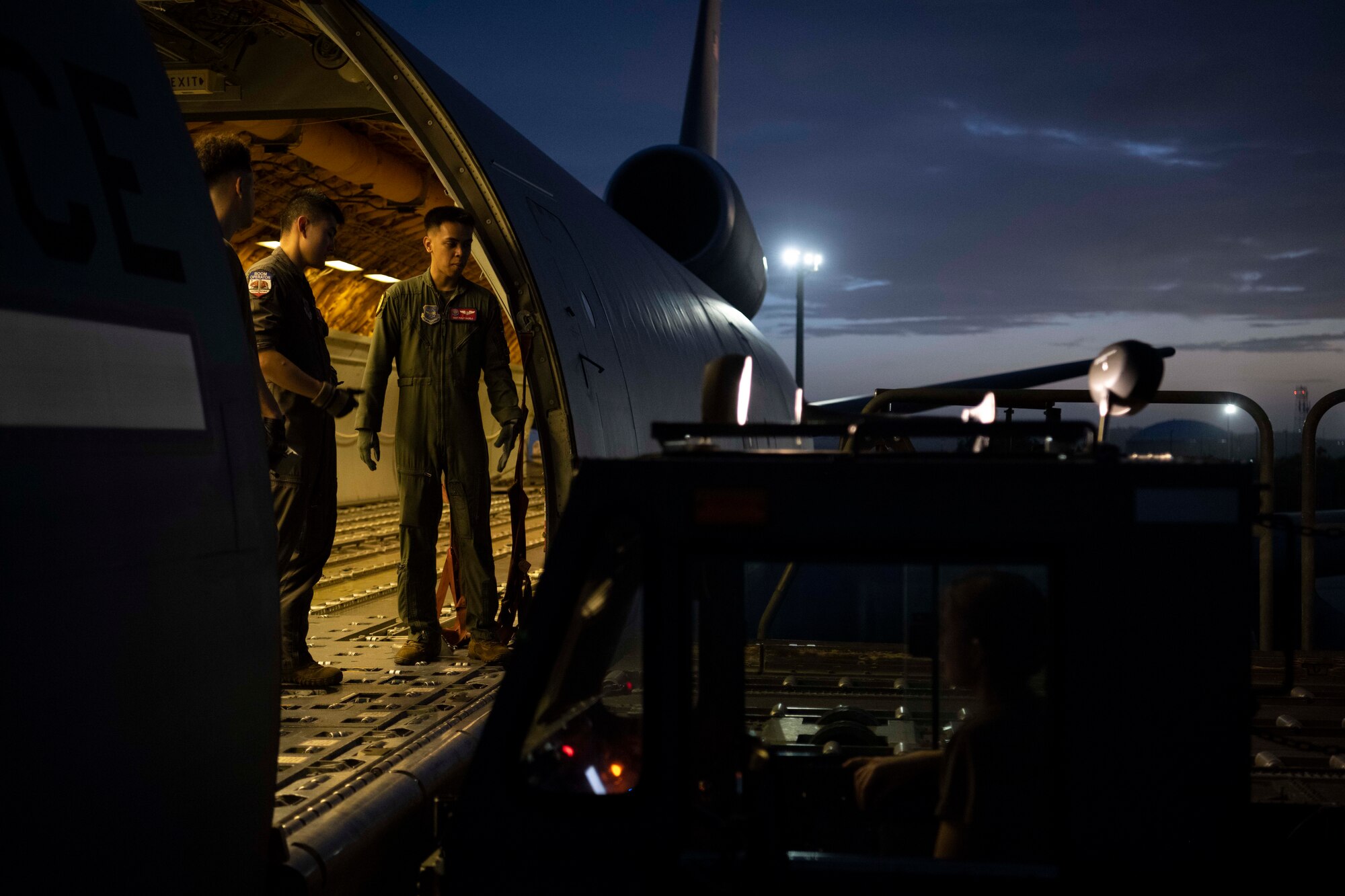 In the dark two military members stand at the loading door of a large military aircraft. A pallet is being loaded from a large military transport vehicle on to the aircraft.