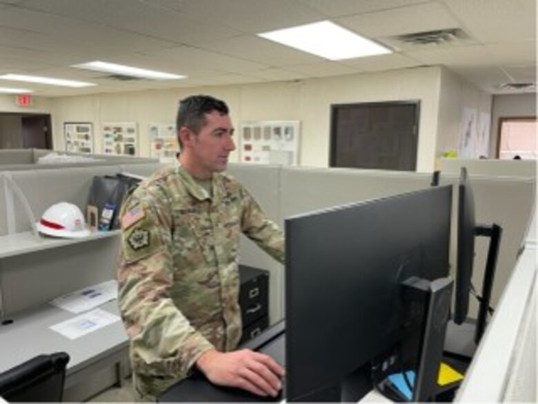 Chief Warrant Officer 2 Adam Isdale stands at his desk and reviews content in a U.S. Army uniform.