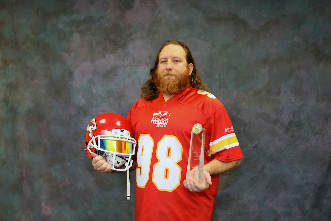 Man in red jersey poses with glass trophy and red football helmet with a gray background.