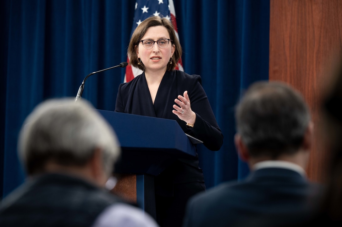 A briefer gestures while speaking to reporters from a lectern with an American flag in the background.