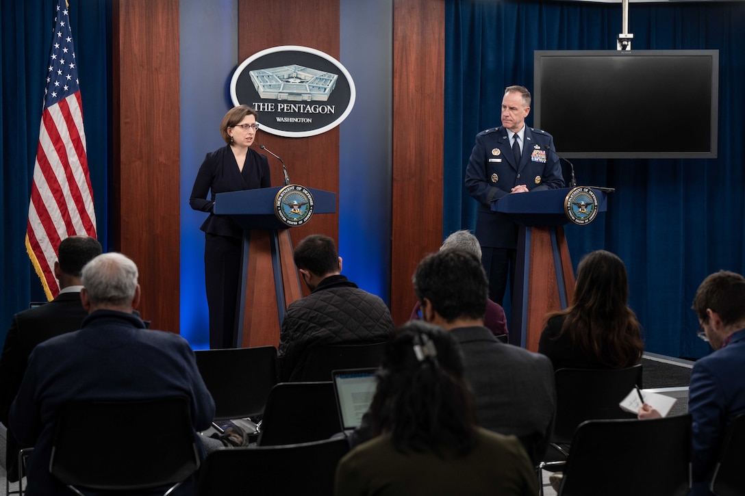 Two briefers, one in uniform speak to reporters from lecterns with the pentagon logo, a tv monitor and American flag in the background.