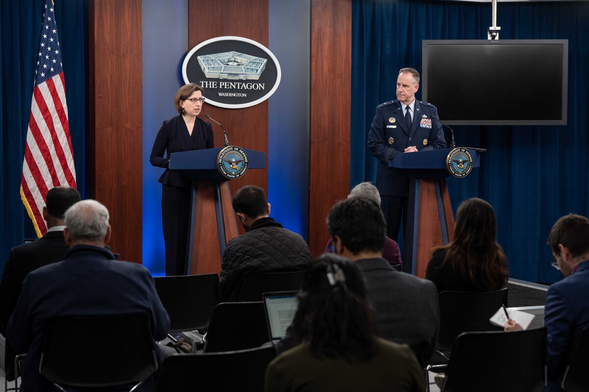 Two briefers, one in uniform speak to reporters from lecterns with the pentagon logo, a tv monitor and American flag in the background.