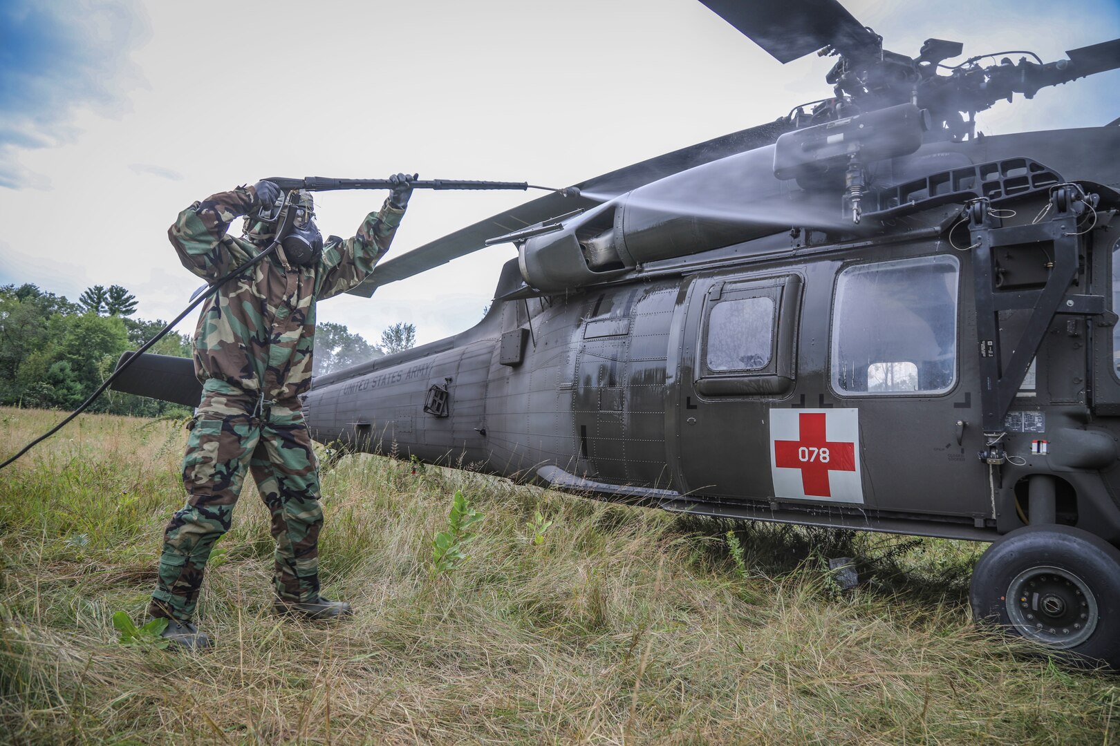 A soldier wearing a respirator mask sprays water from a hose onto a helicopter.