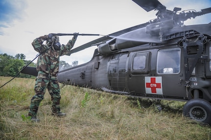 A soldier wearing a respirator mask sprays water from a hose onto a helicopter.