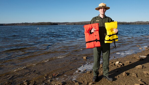 Andrew Harrell stands in front of the beach area at Cook Recreation area holding two life jackets.