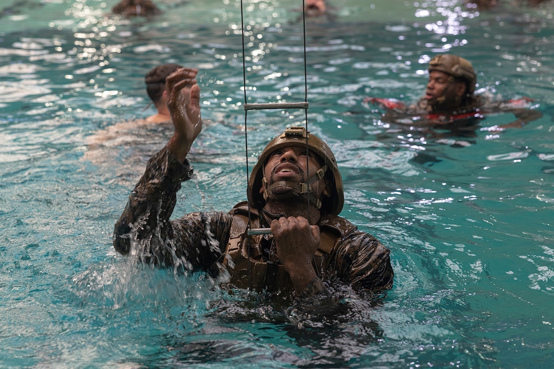 A service member in uniform and helmet is shown reaching for a ladder while in a pool.