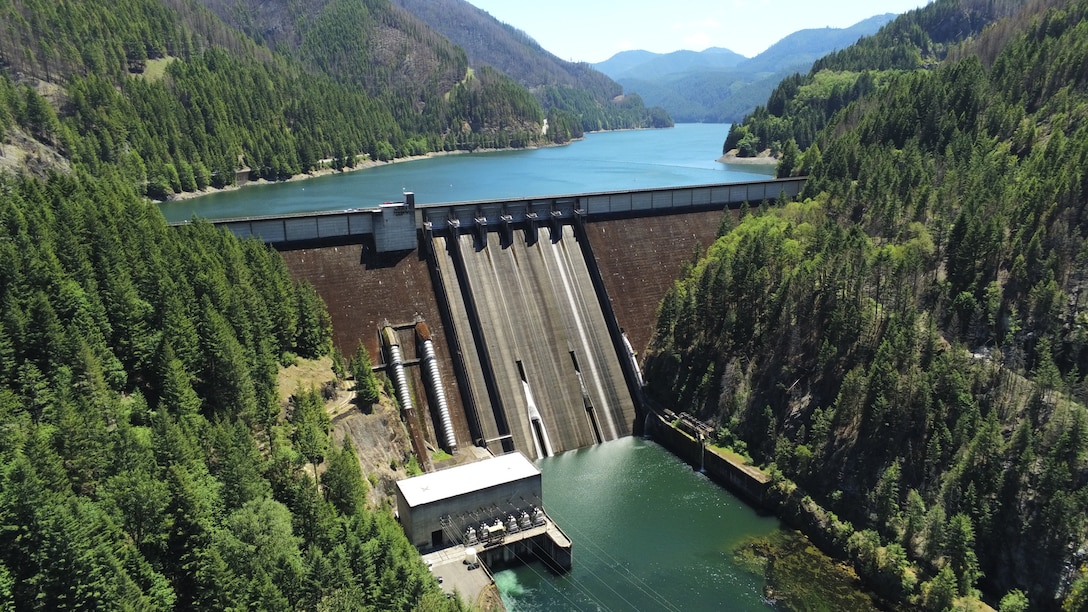 A large concrete dam hundreds of feet high with a lake behind it and evergreen-covered hills around it on a clear, sunny summer day.