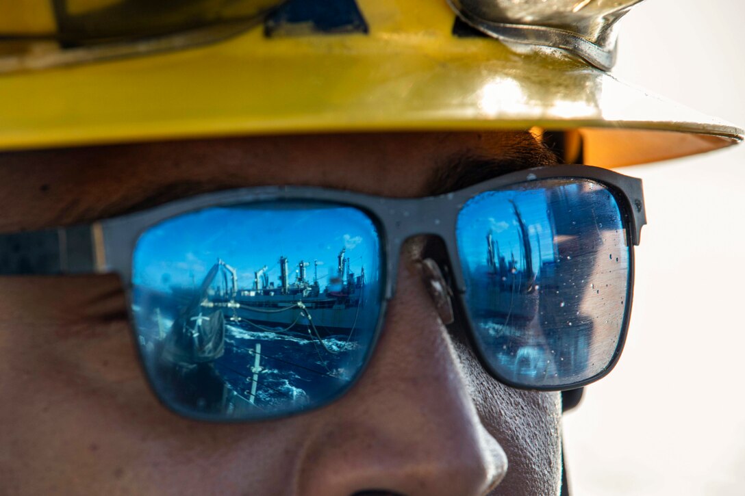 A ship sails next to another ship connected by a hose as seen through the reflection of a sailor’s sunglasses.