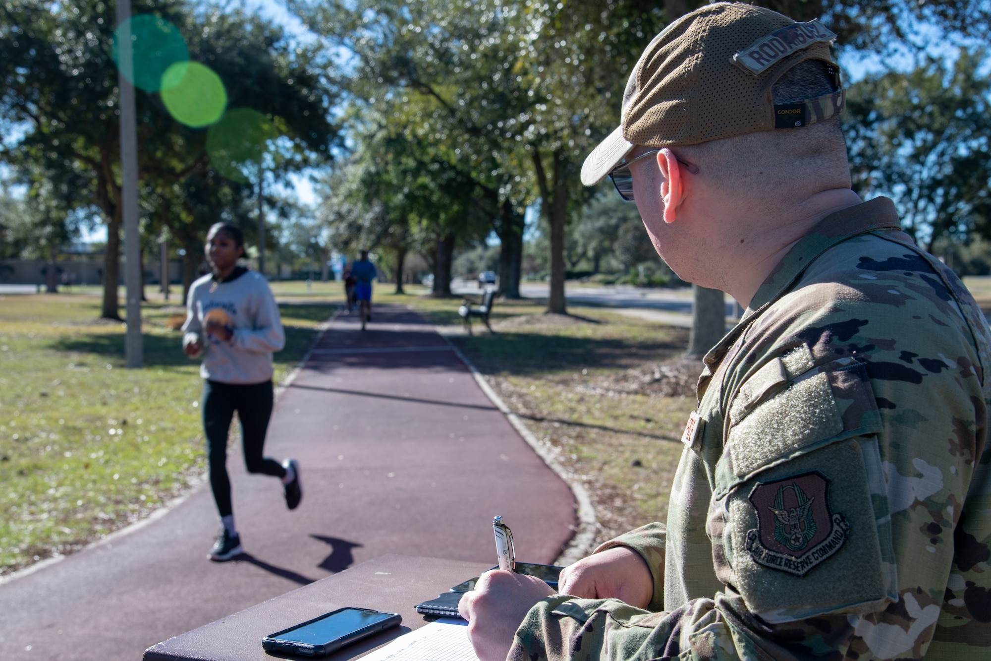 A person runs on a track in the background as MSgt Rodriguez marks a sheet of paper while looking at the runner