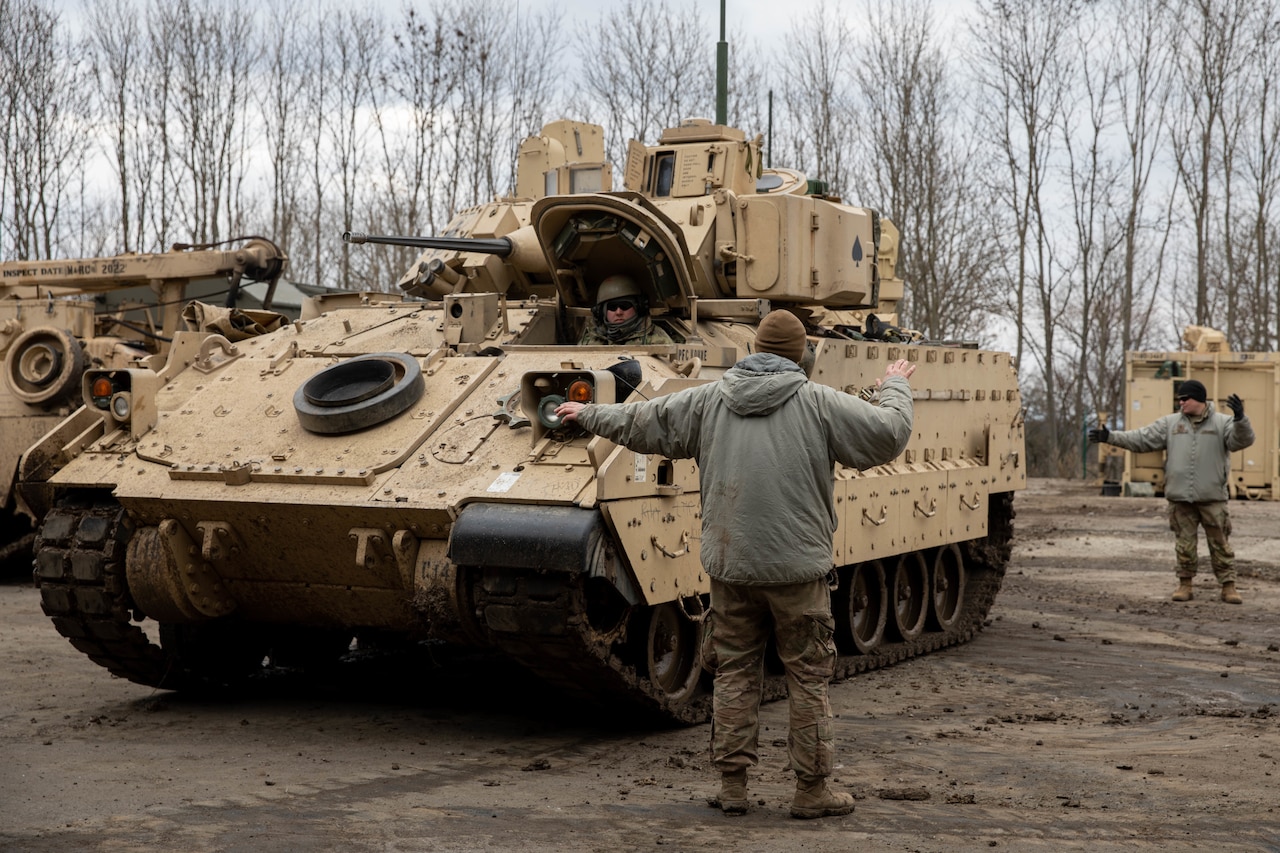 A soldier looks out the hatch of an armored military vehicle as one soldier in front of him and another in the rear use hand gestures to guide the vehicle’s movements.