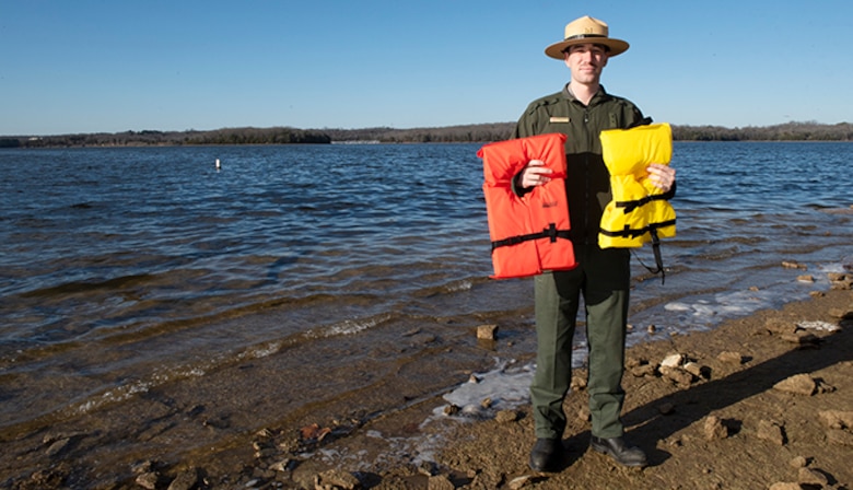 Andrew Harrell stands in front of the beach area at Cook Recreation area holding two life jackets.