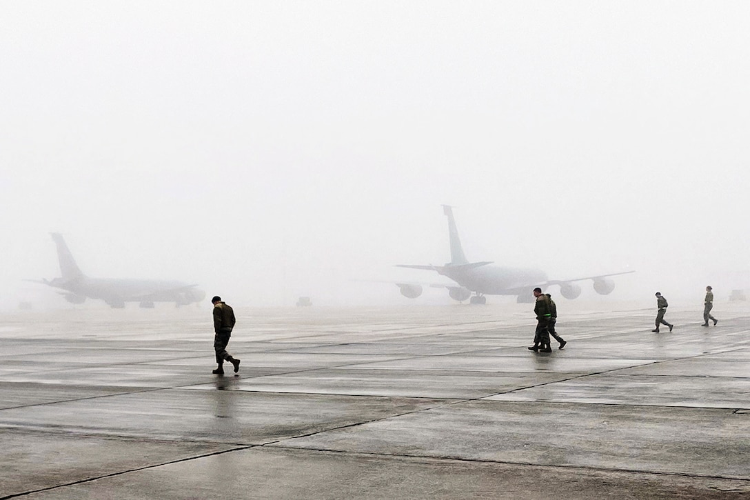 Airmen search for trash and objects on the flight line as fog sets in with military aircraft behind them.