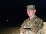 White male adult in US Army camouflage uniform stands looking at the camera with his arms crossed. He is front lit at night, with a dark background. The photo is from waste up. He is wearing sergeant rank on his jacket and cap.