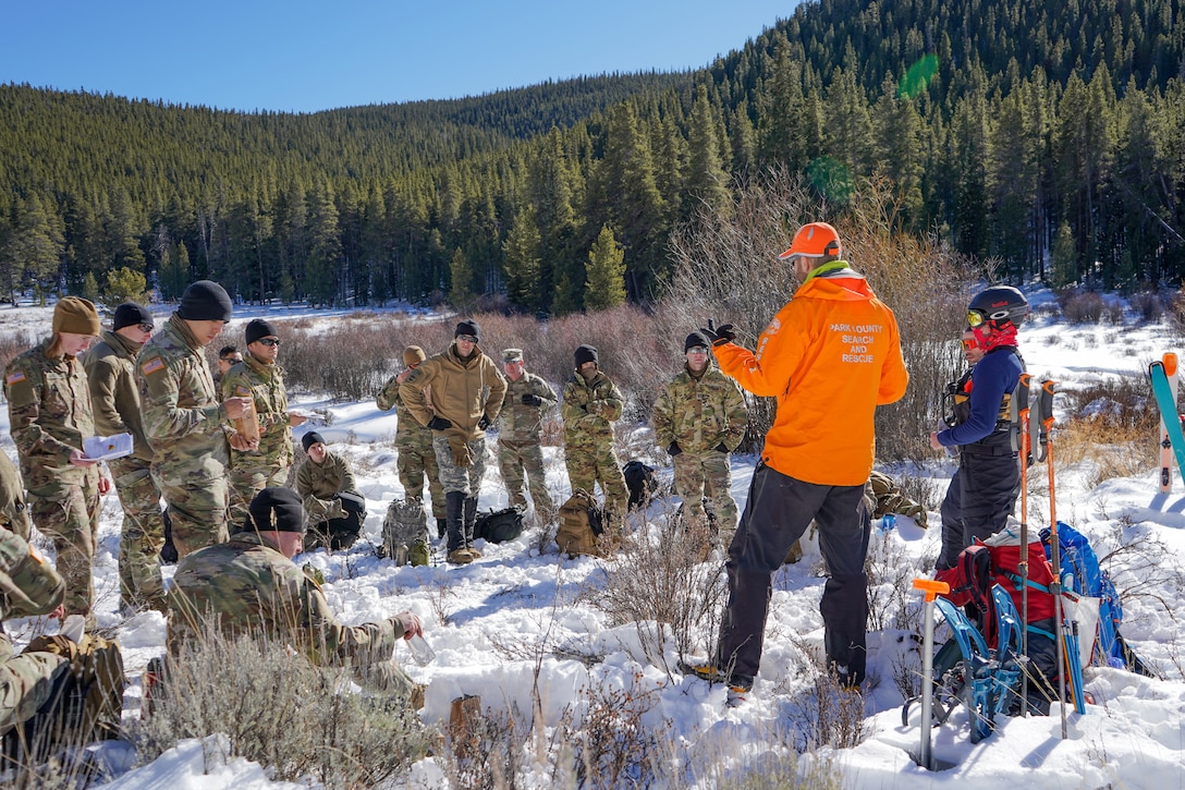 A group of soldiers listen to an outdoor lecture in a wooded environment.