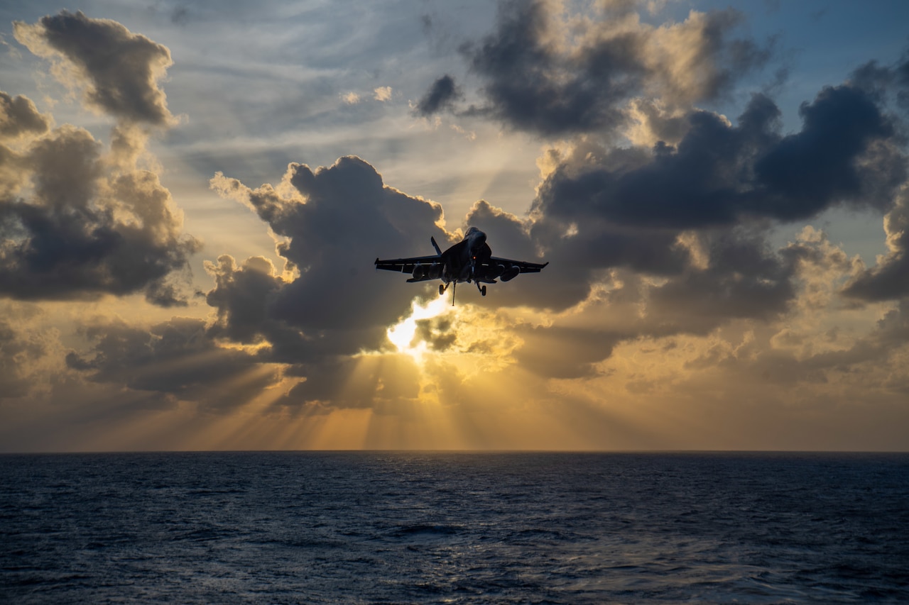A fighter jet comes in for a landing on an aircraft carrier at twilight.