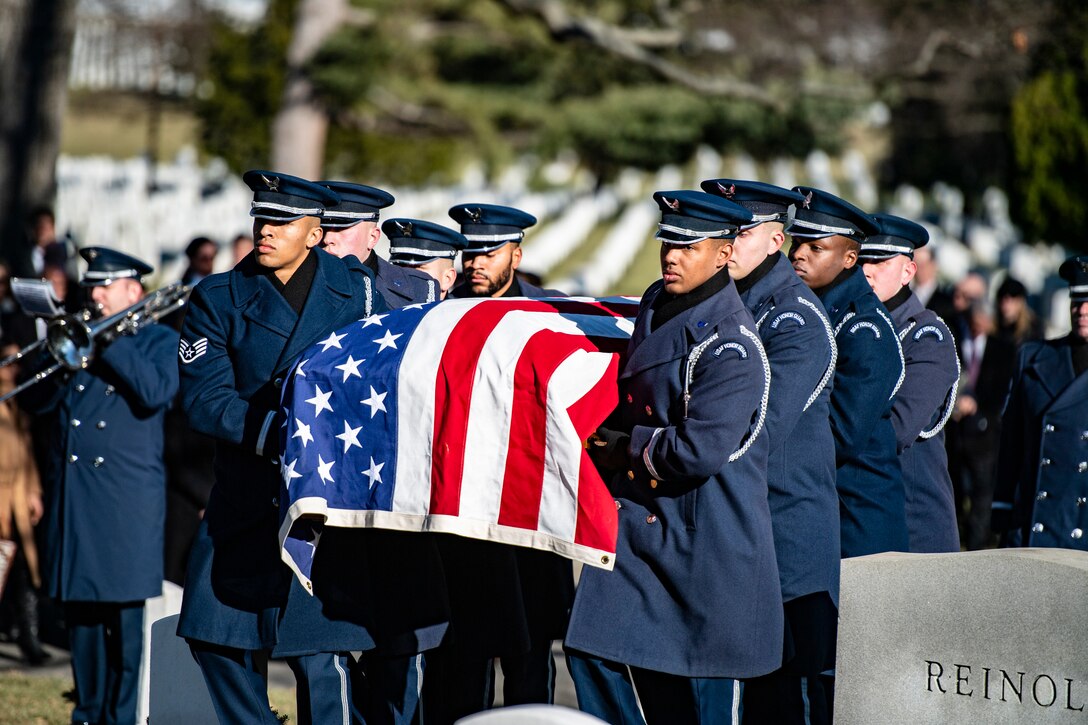 Service members carry a flag-draped casket in a cemetery.