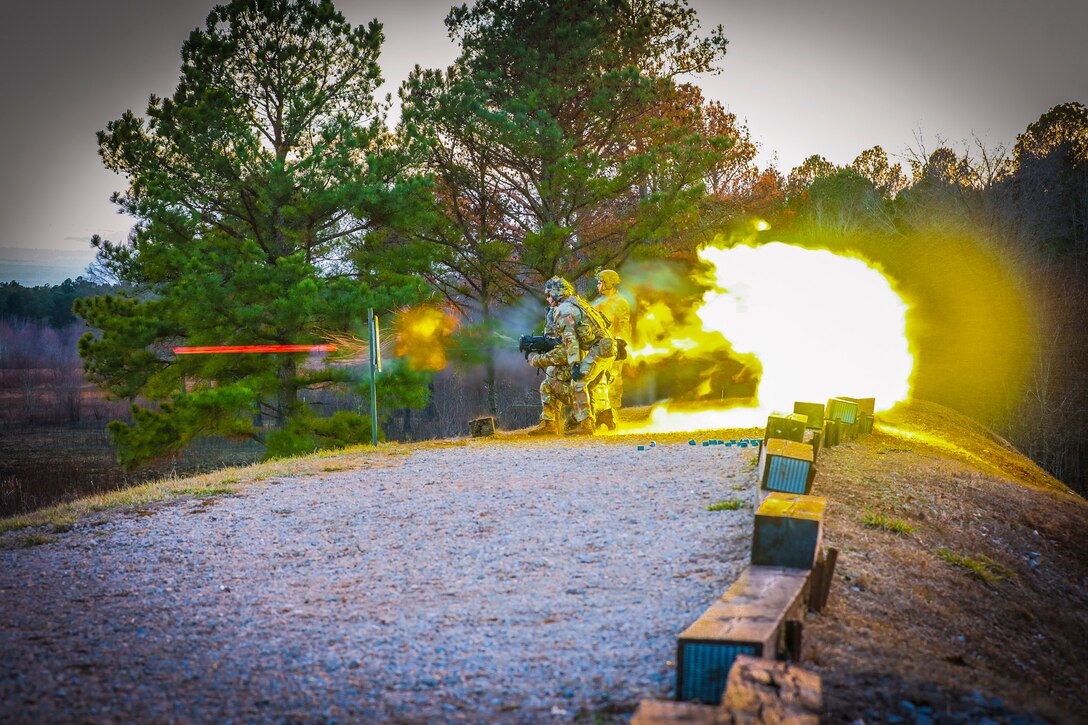 Flames shoot from the back of a weapon that soldiers fire at an outdoor range.