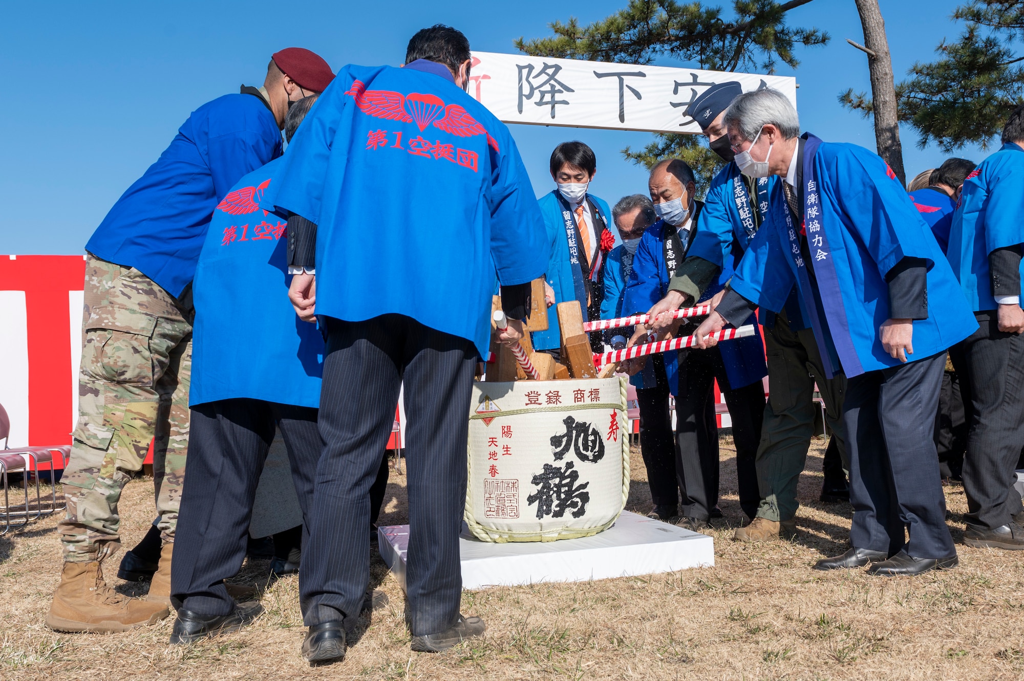A dozen people holding wooden mallets break open a sake barrel