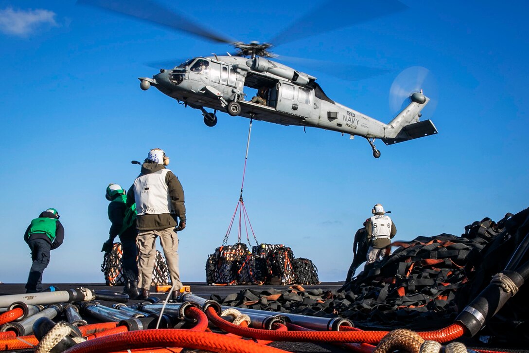 A helicopter delivers supplies to the deck of a ship while sailors watch